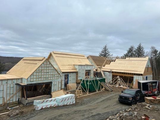 Maison en construction, murs et toit inachevés avec poutres en bois apparentes, ciel nuageux.