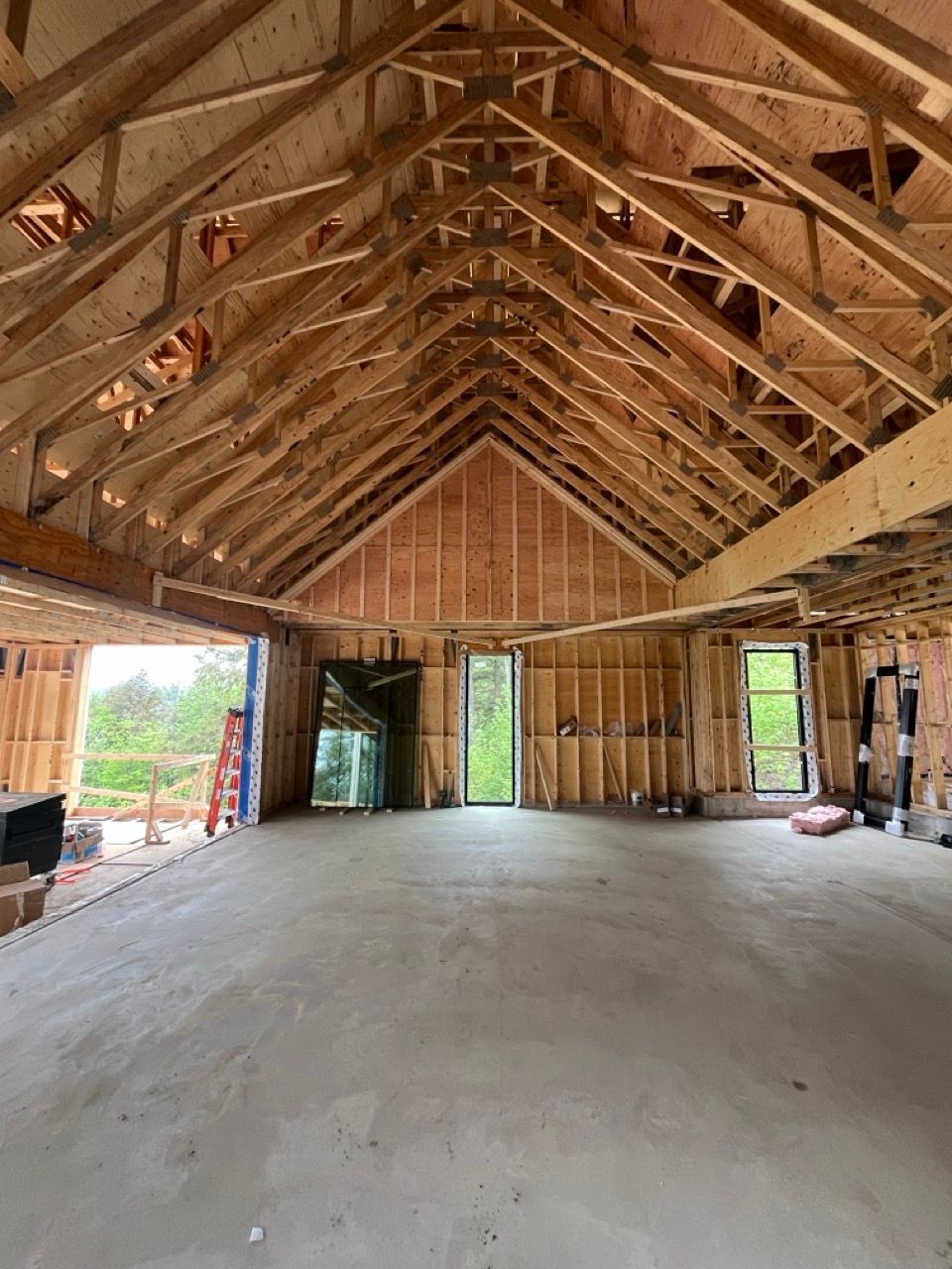 Vue intérieure d'un bâtiment en construction, montrant la charpente en bois du toit et des murs, le sol en béton et les fenêtres.
