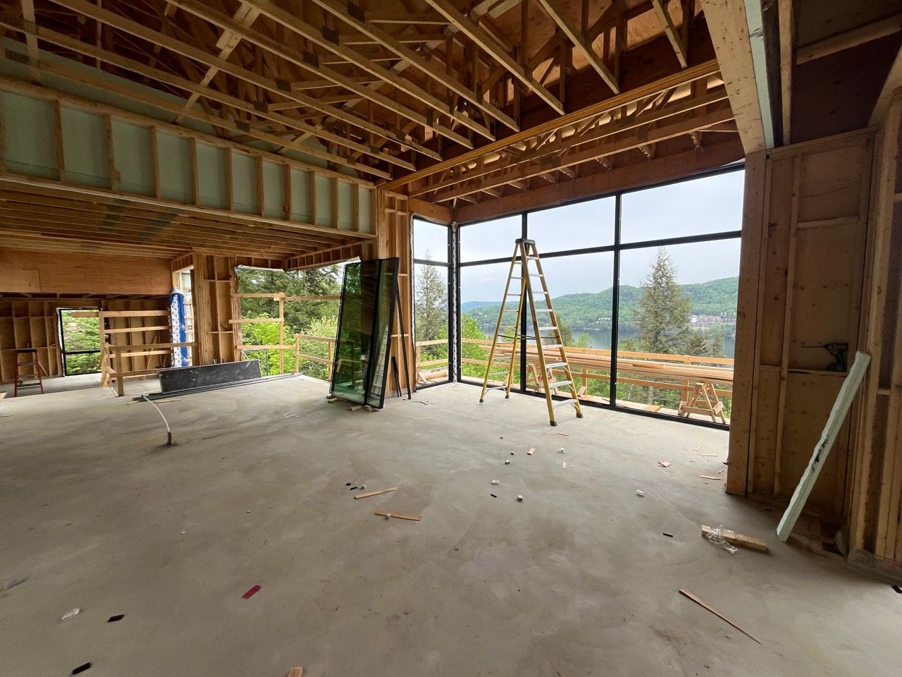 Intérieur d'une maison en construction avec une charpente en bois apparente et de grandes fenêtres donnant sur une colline verdoyante.
