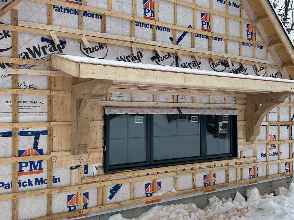 Extérieur d'une maison en construction ; charpente en bois, fenêtre et légère accumulation de neige.