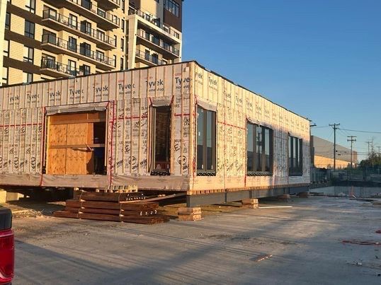 Bâtiment modulaire en construction sur une zone pavée, avec fenêtres et bardage en contreplaqué, à proximité d'un bâtiment plus haut.