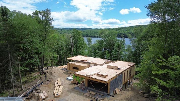 Construction d'une maison moderne dans une clairière, avec un lac en arrière-plan sous un ciel bleu.