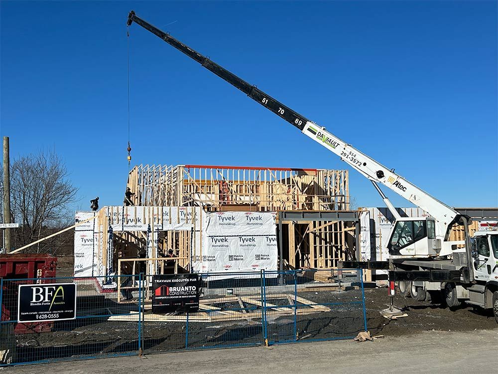 Chantier de construction avec une grue soulevant des matériaux pour construire une structure à ossature bois sur fond de ciel bleu.