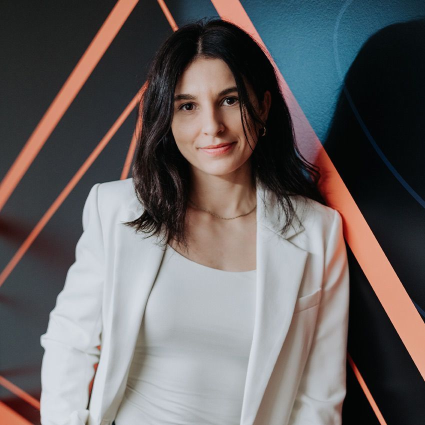 Femme en blazer et haut blancs, souriante, appuyée contre un mur orné d'un motif abstrait orange et bleu.