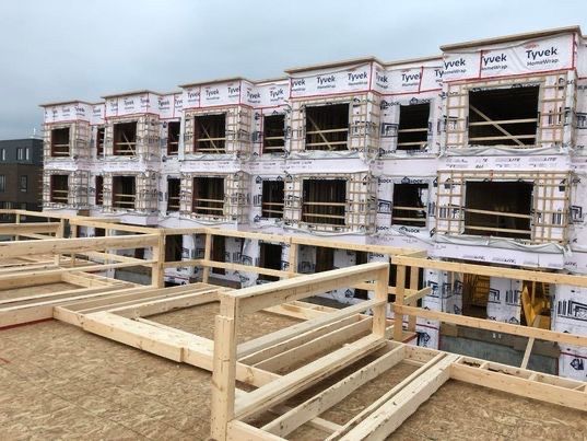 Immeuble à plusieurs étages en construction, avec charpente en bois apparente, fenêtres et garde-corps de sécurité.