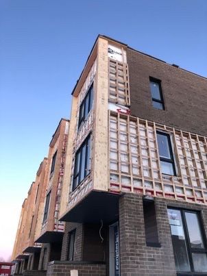 Immeuble à plusieurs étages en construction ; structure en bois apparente, briques sombres et fenêtres sur fond de ciel bleu.