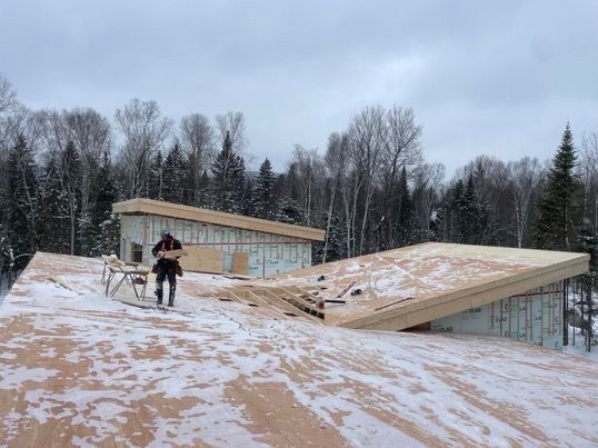 Construction d'une maison moderne dans une forêt enneigée. Un ouvrier marche sur un toit en bois.