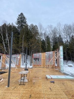 Chantier de construction : structure en bois d'un bâtiment avec isolation, charpente et poteau métallique, située dans une zone boisée enneigée.