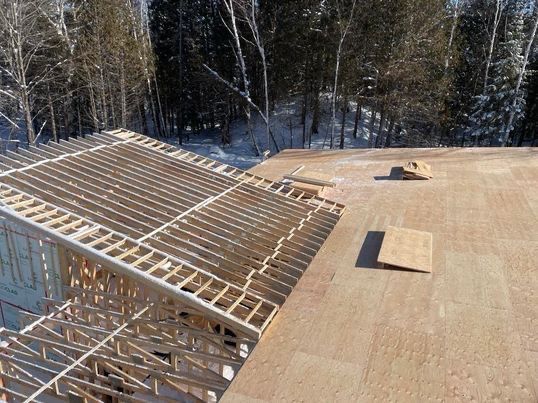 Construction de la charpente d'un toit, montrant les poutres en bois et le contreplaqué apparents, sur fond de forêt hivernale.