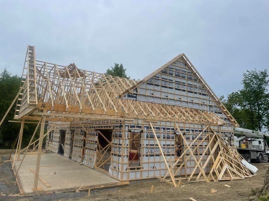 Maison en construction : ossature bois avec toit à pignon et bardage, porche en béton. Ciel nuageux.