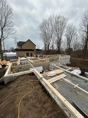 Chantier de construction ; charpente en bois sur fondations en béton ; une maison à deux étages en arrière-plan.