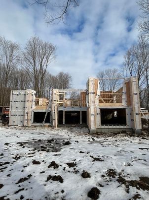 Chantier de construction d'une maison à ossature bois et panneaux isolants, situé dans une zone boisée et enneigée sous un ciel nuageux.