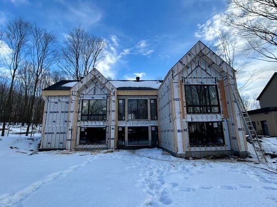 Maison en construction dans un décor enneigé, avec charpente apparente et grandes fenêtres sous un ciel bleu.