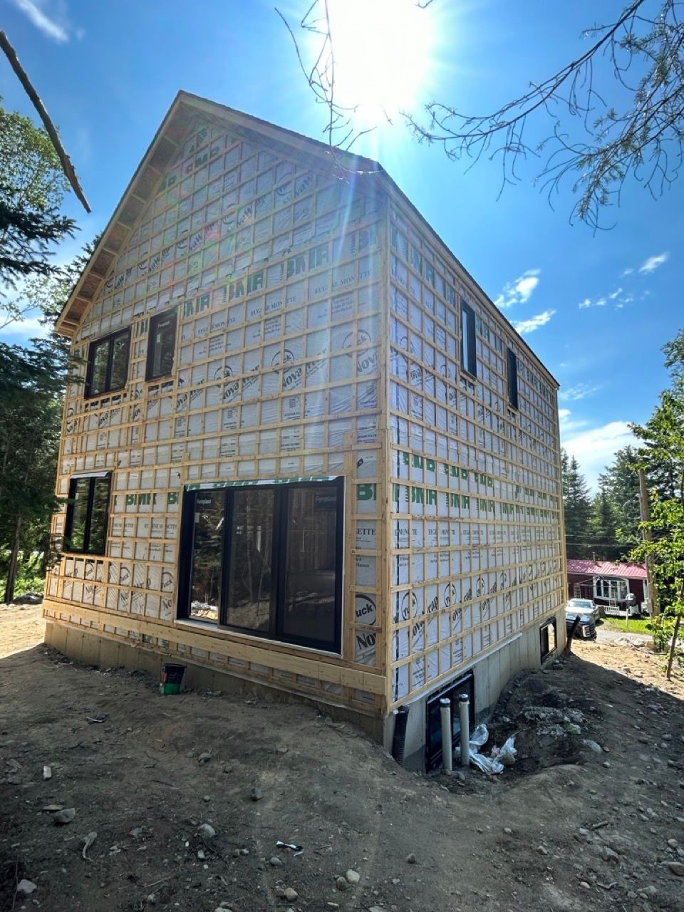 Maison à deux étages en construction, avec fenêtres et bardage, située sur un terrain légèrement en pente par une journée ensoleillée.