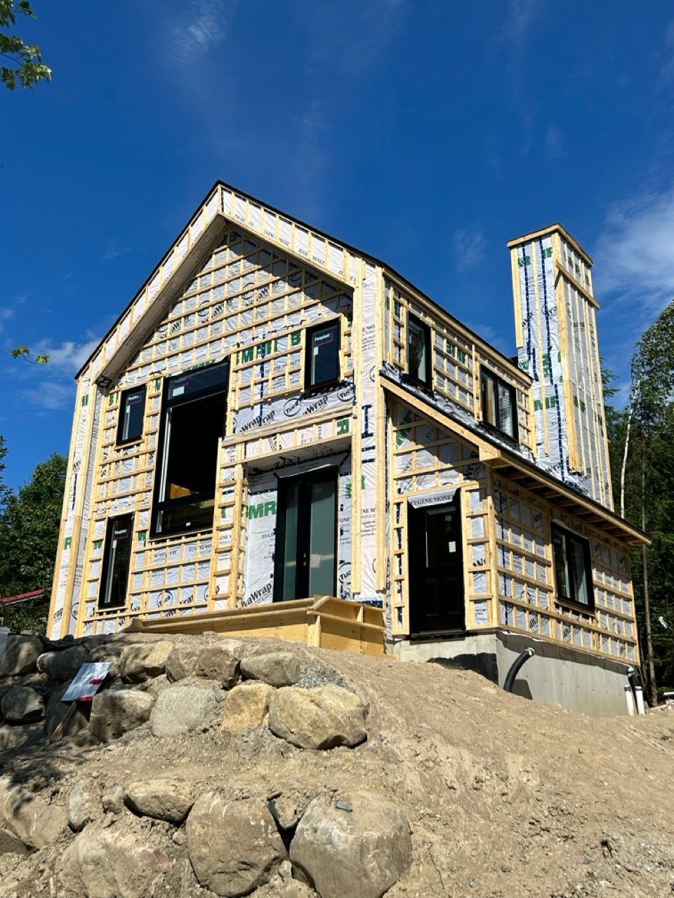 Maison à deux étages en construction. Ossature bois clair, encadrements de fenêtres noirs, cheminée et ciel bleu.