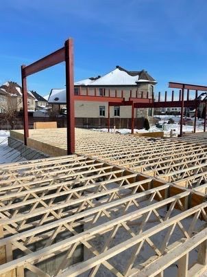 Chantier : solives en bois, poutres d'acier rouges, neige, ciel bleu, maisons en arrière-plan.