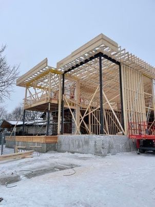Chantier de construction : ossature en bois d’un bâtiment à deux étages sur fondations en béton, avec une poutre de soutien noire ; sol enneigé.