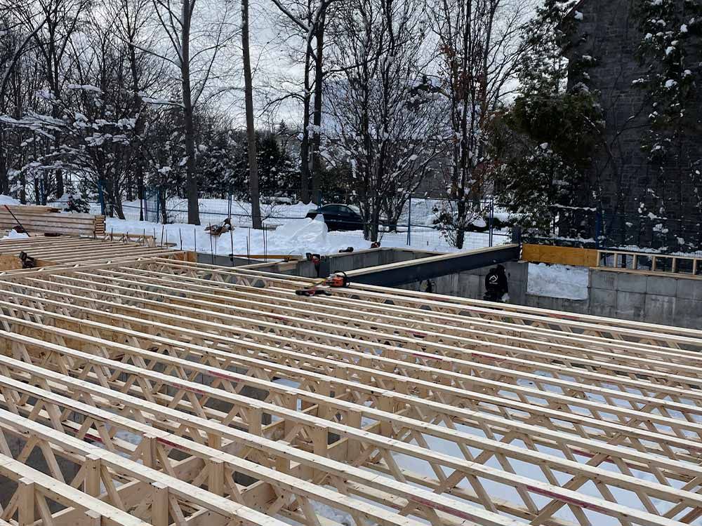 Chantier de construction avec charpentes en bois posées sur un sol enneigé. Des ouvriers sont visibles, dans un paysage hivernal.