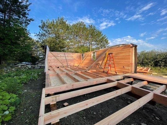 Bâtiment en bois en construction ; sol et murs non finis, échelle. Extérieur avec arbres et ciel bleu.