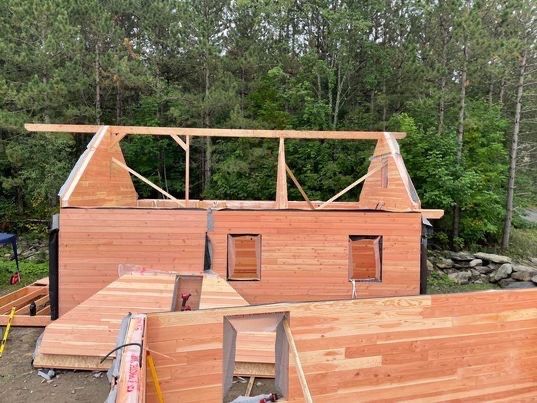 Structure à ossature bois en construction ; bardage en cèdre, toit ouvert, arbres en arrière-plan.