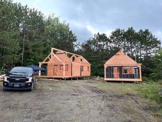 Deux charpentes de cabanes en bois sont en construction dans une zone boisée, avec une camionnette garée à proximité.