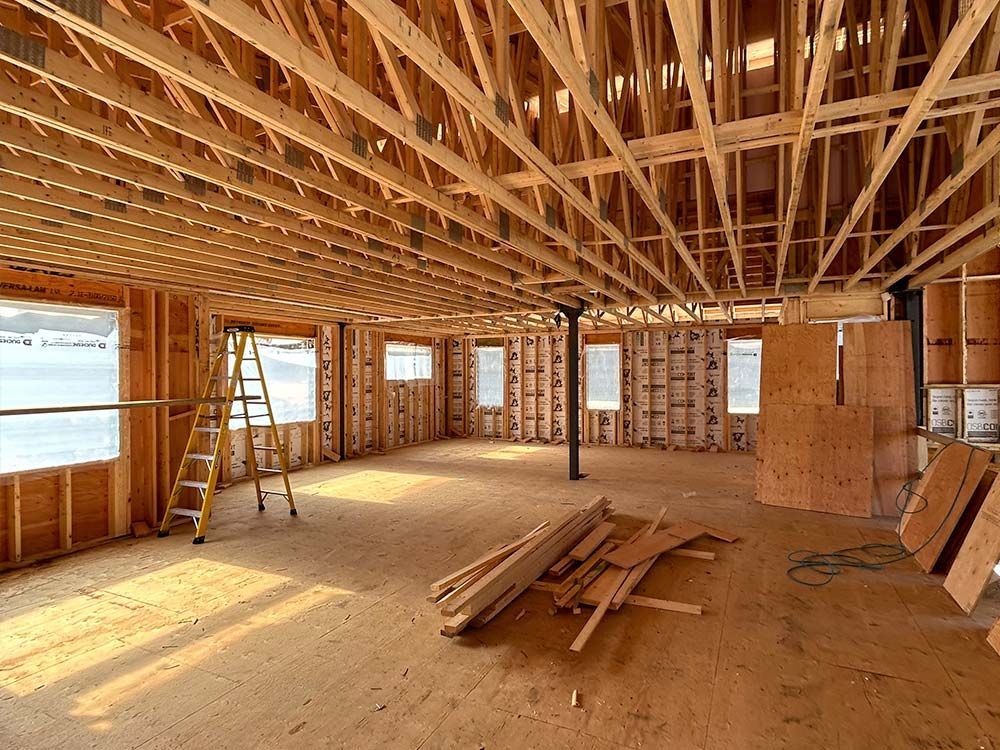 Intérieur d'une maison en construction ; charpente et chevrons en bois apparents, fenêtres, échelle et débris de bois au sol.