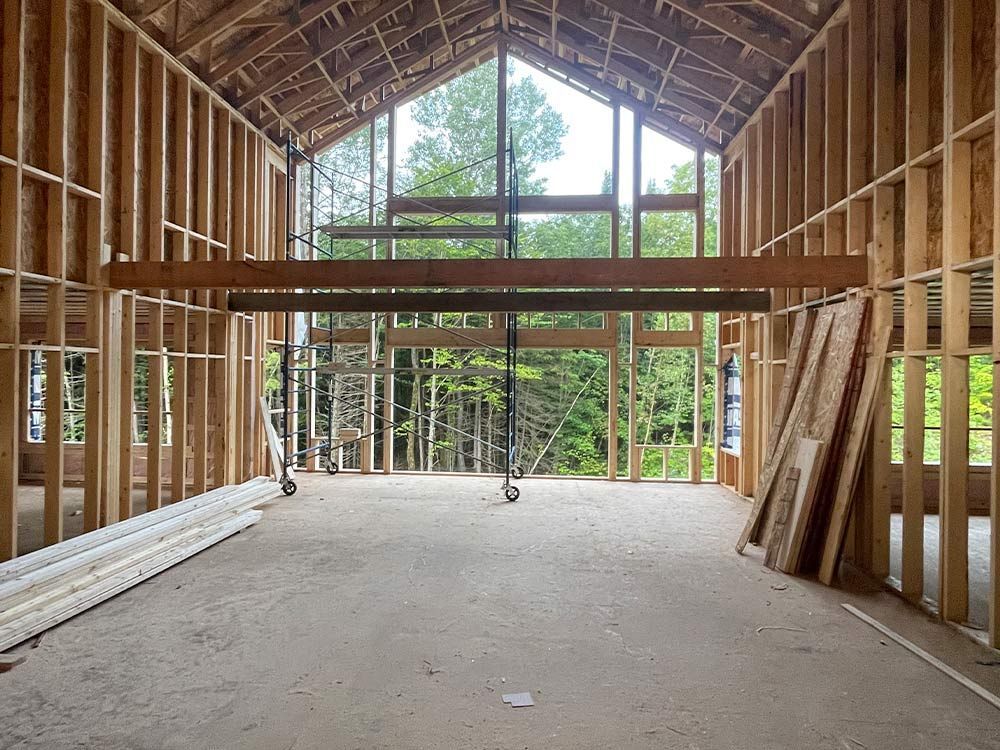 Vue intérieure d'un bâtiment en construction, à ossature de poutres en bois. Les encadrements de fenêtres laissent entrevoir des arbres.