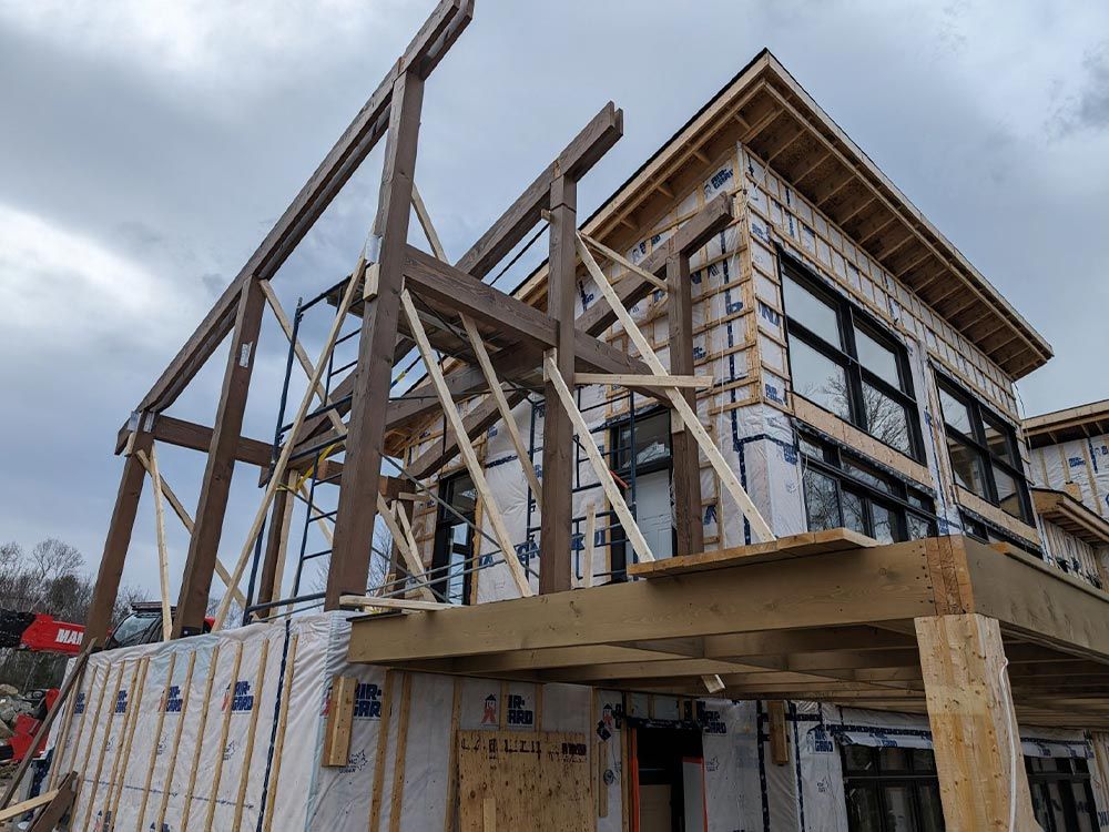 Chantier de construction d'une maison avec charpente en bois apparente, bardage et grandes fenêtres sous un ciel nuageux.