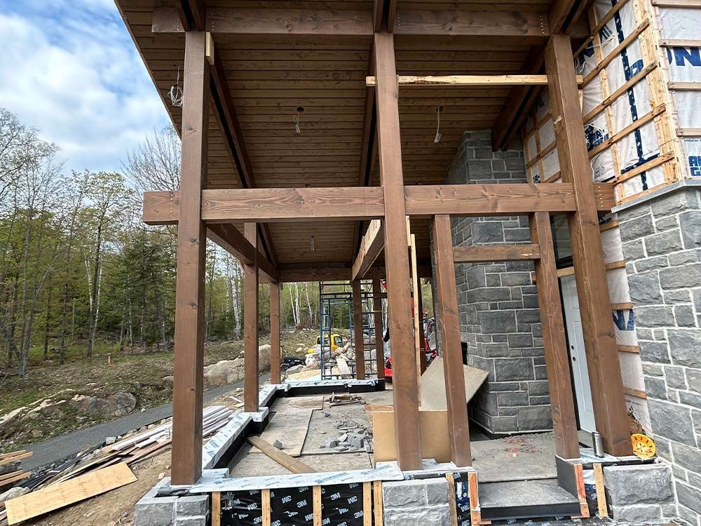 Porche en bois en construction ; poutres brunes, cheminée en pierre grise, ciel bleu.