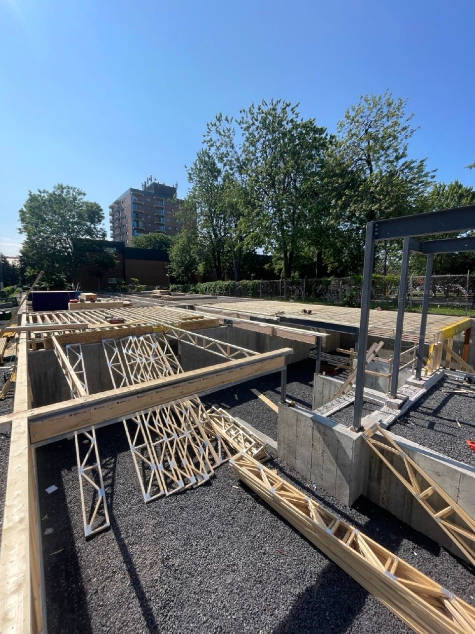 Chantier de construction avec charpente en bois et fondations en béton sur gravier. Ciel bleu vif, arbres.