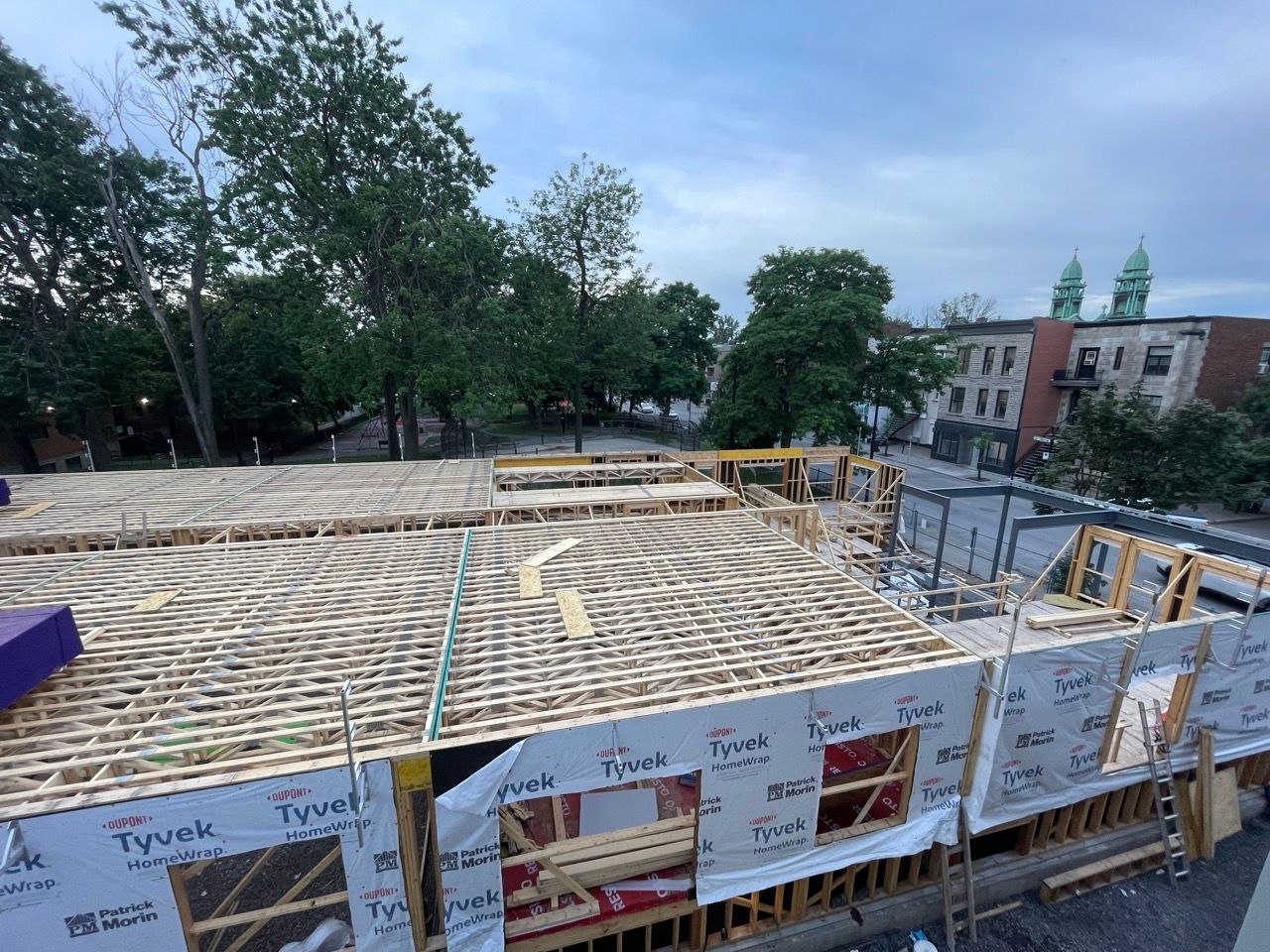 Chantier de construction avec charpente en bois apparente et bâtiments environnants sous un ciel nuageux.