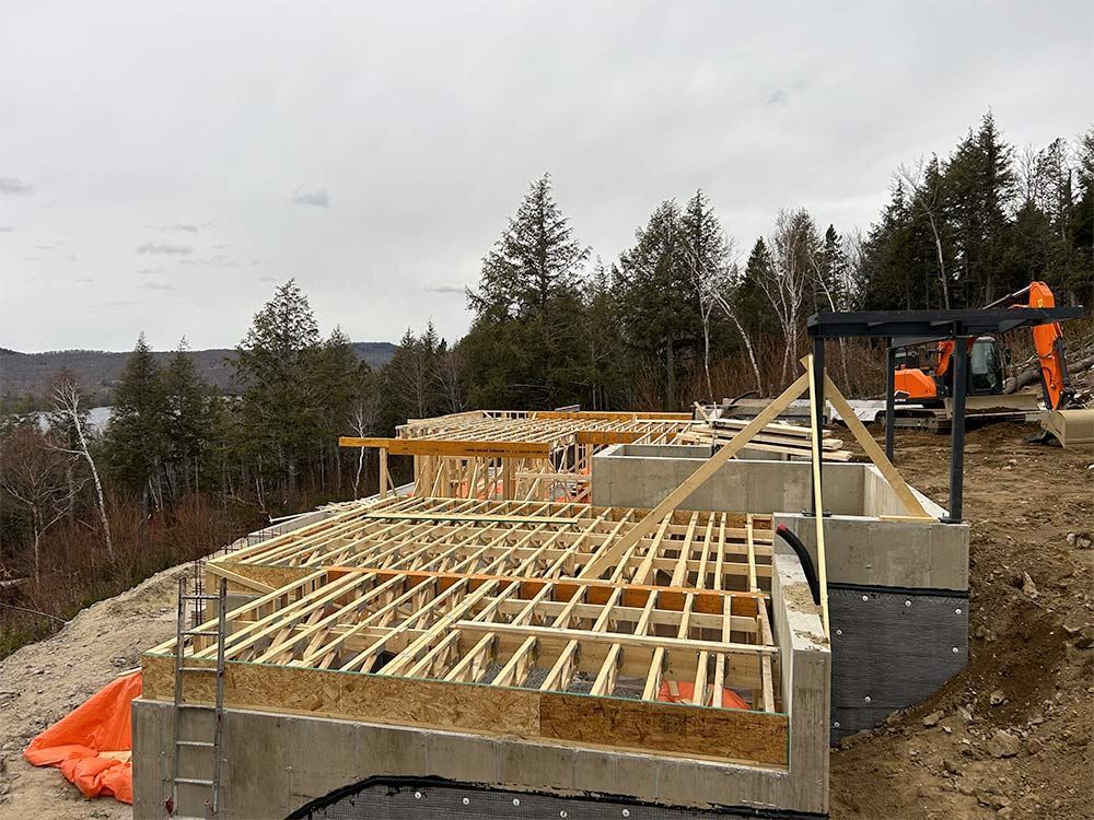 Chantier : structure en bois sur fondations en béton, surplombant un lac et des arbres sous un ciel nuageux.
