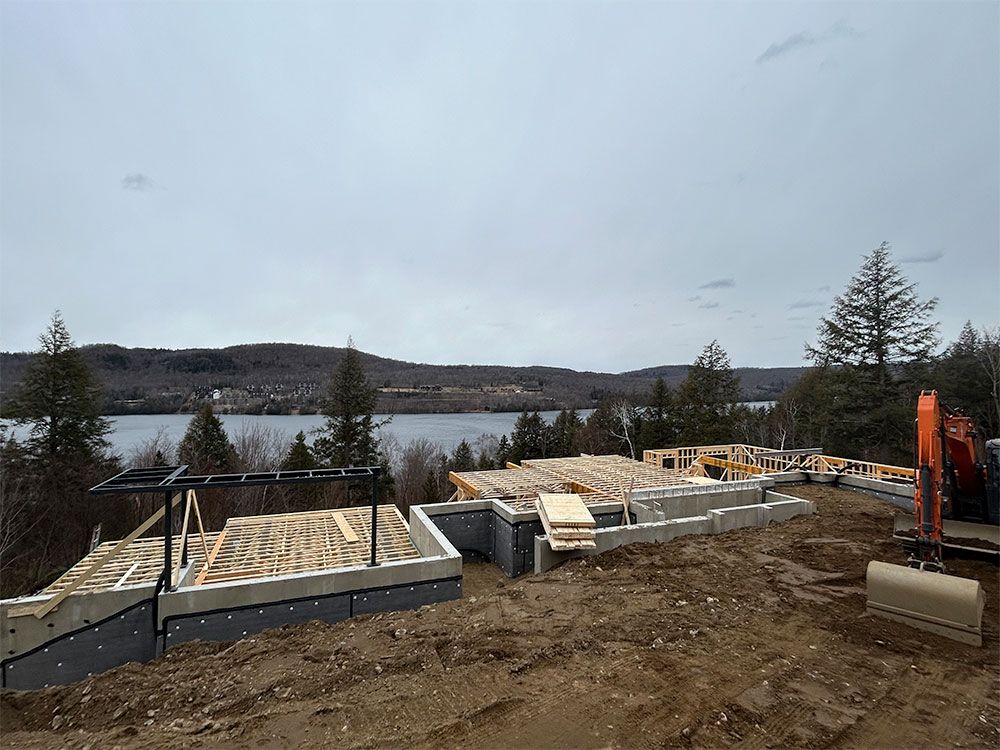 Chantier de construction avec fondations, charpente en bois et excavatrice donnant sur un lac et des collines.