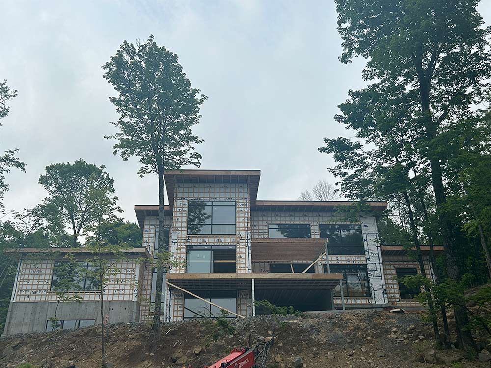 Maison moderne en construction, entourée d'arbres. Grandes fenêtres, murs en parpaings clairs, ciel couvert.