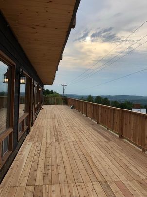 Terrasse en bois avec rambarde longeant un bâtiment aux grandes fenêtres. Ciel couvert avec vue au loin.