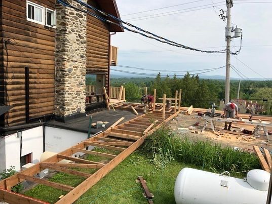 Construction d'une terrasse et d'une rampe en bois attenantes à une maison en rondins. Des personnes travaillent sur la structure.