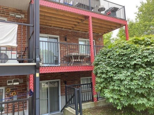 Balcons d'appartements à cadre rouge, attenants à un immeuble en briques. Garde-corps noirs et portes coulissantes en verre. Buisson vert à droite.