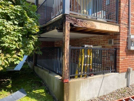 Balcon endommagé avec bois pourri et structure apparente, jouxtant un bâtiment en briques.