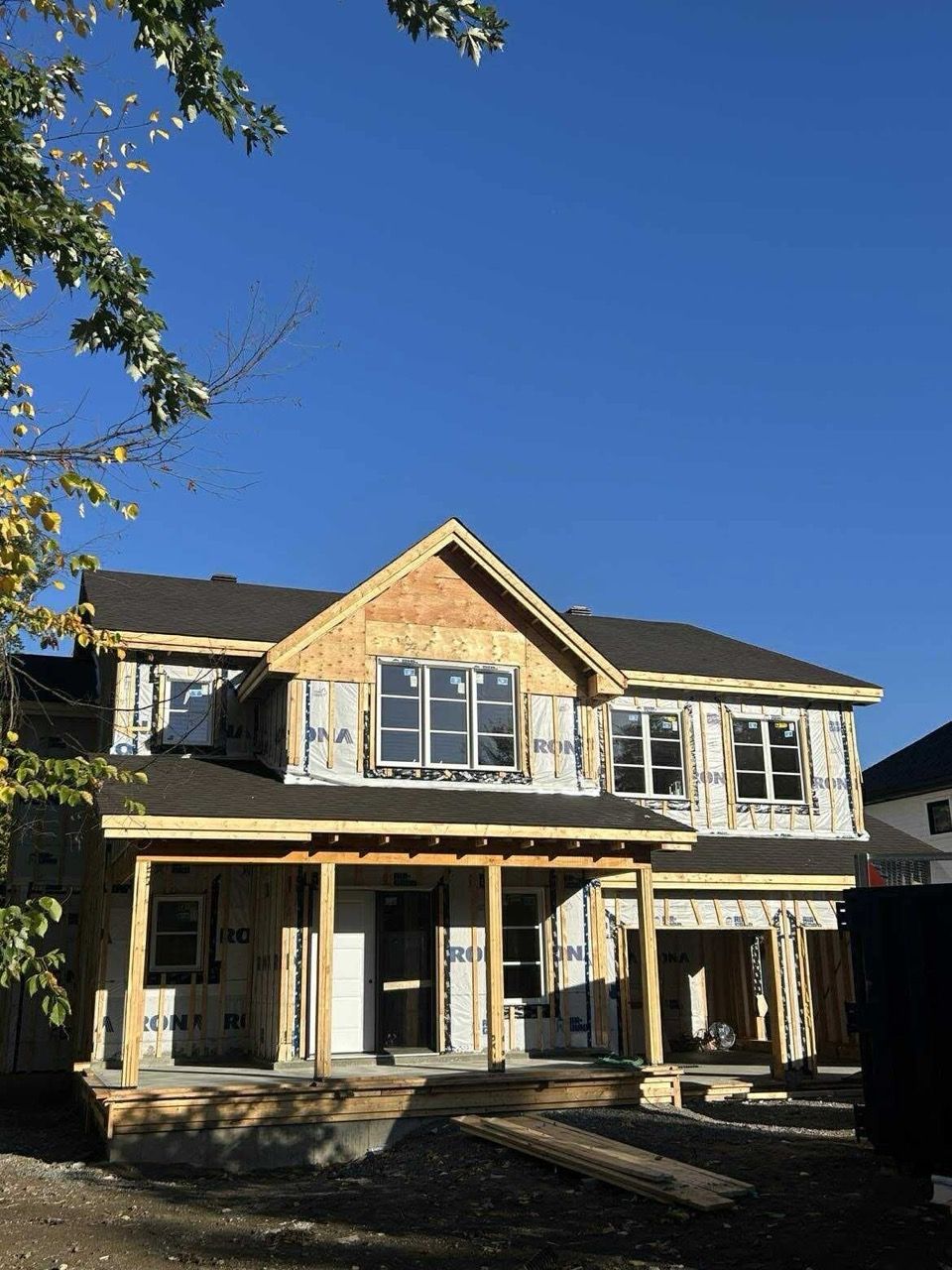 Maison à deux étages en construction, avec un ciel bleu en arrière-plan. Charpente en bois apparente visible, y compris le porche et le pignon supérieur.