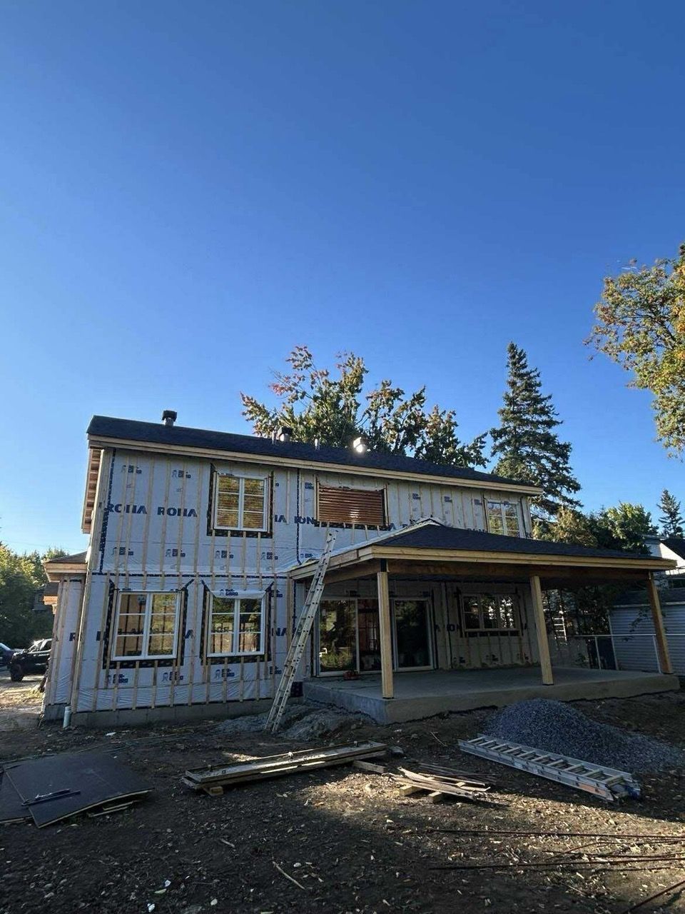 Maison à deux étages en construction, recouverte d'un film bleu. Un porche couvert et les encadrements de fenêtres se détachent sur le ciel bleu.