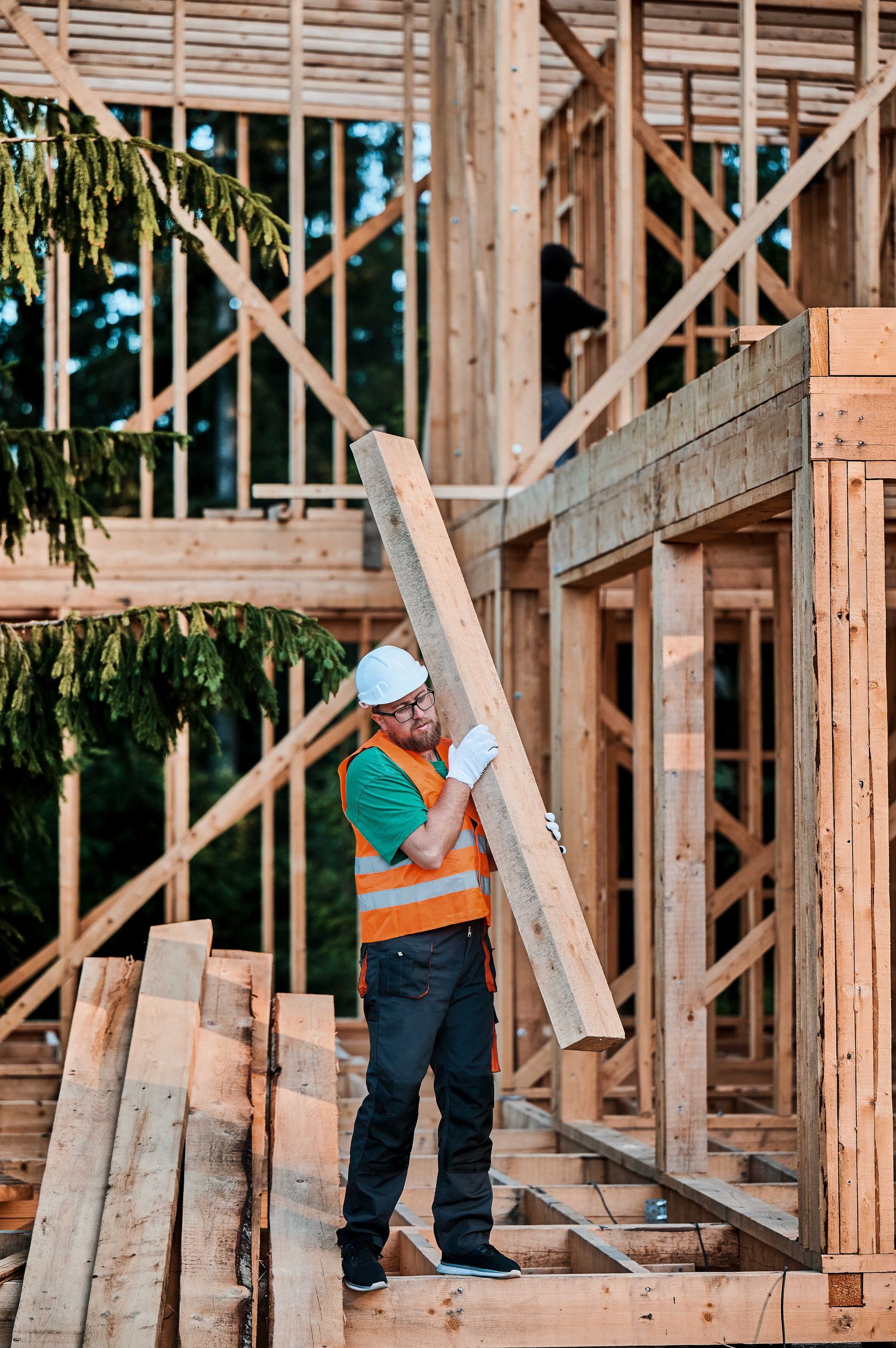 Ouvrier du bâtiment transportant du bois sur un chantier. Il porte un casque et un gilet de sécurité. Une structure en bois est visible en arrière-plan.