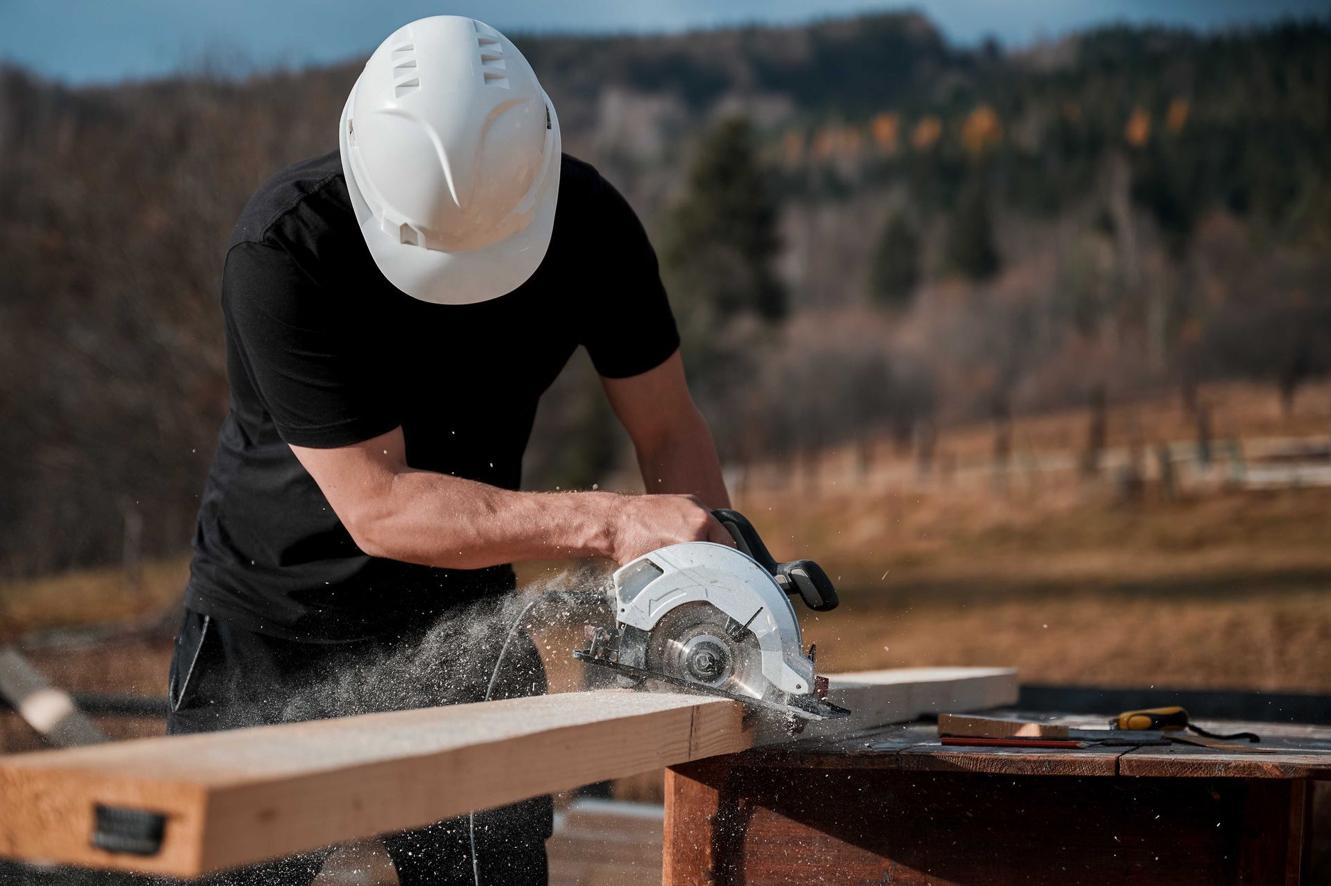 Une personne portant un casque de chantier utilise une scie circulaire pour couper une planche de bois à l'extérieur. De la sciure vole.