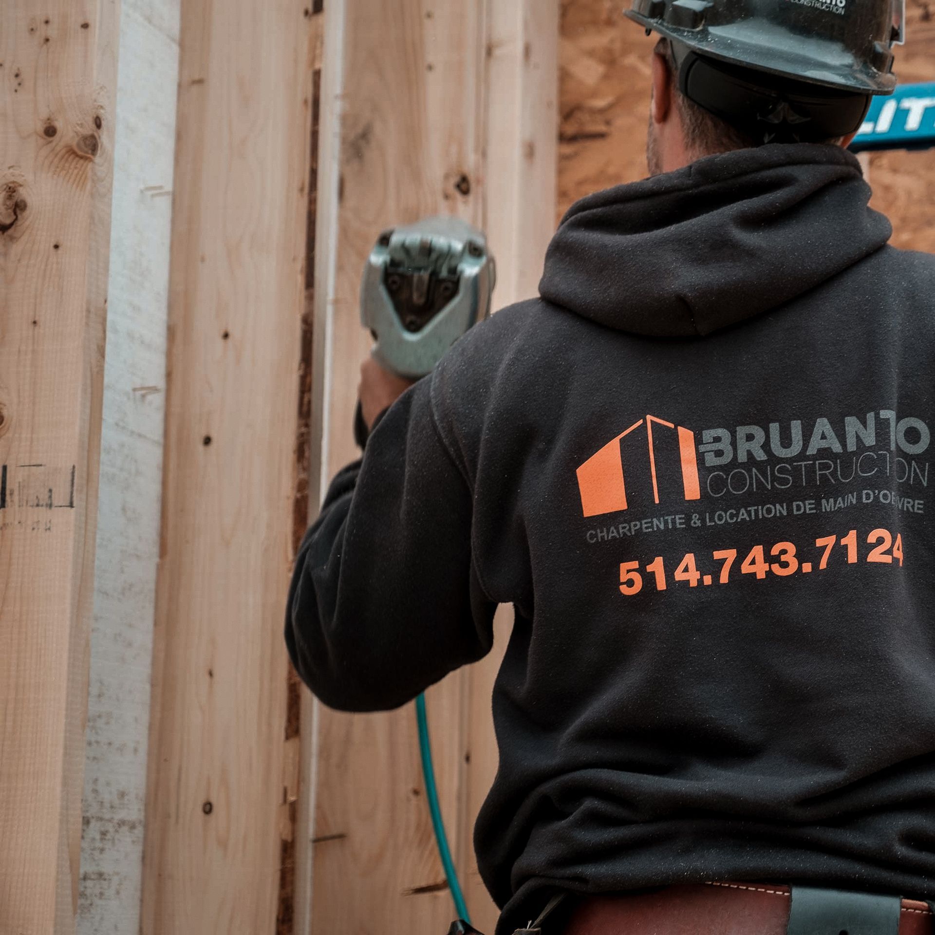 Un ouvrier du bâtiment utilise un cloueur pneumatique sur un mur en bois. Il porte un casque de chantier et un sweat-shirt à capuche avec le logo de son entreprise.