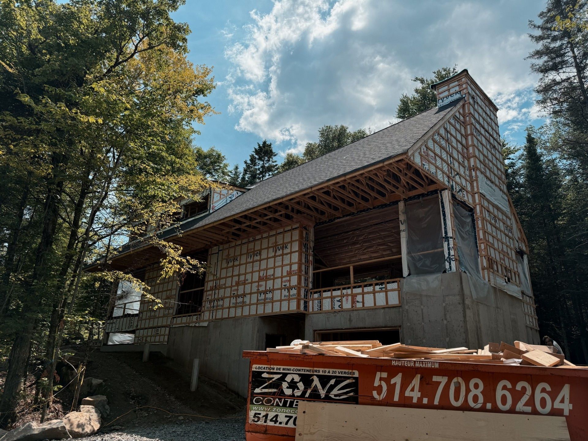 Maison en construction avec charpente apparente, bardeaux bruns et une benne à ordures.