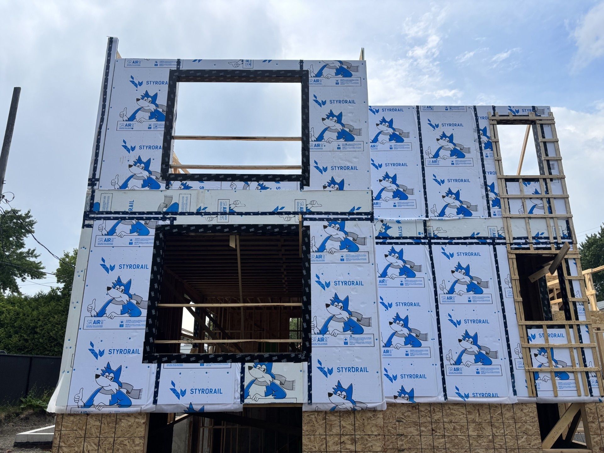 Maison en construction, revêtue d'un matériau bleu et blanc, fenêtres encadrées, se détachant sur un ciel nuageux.