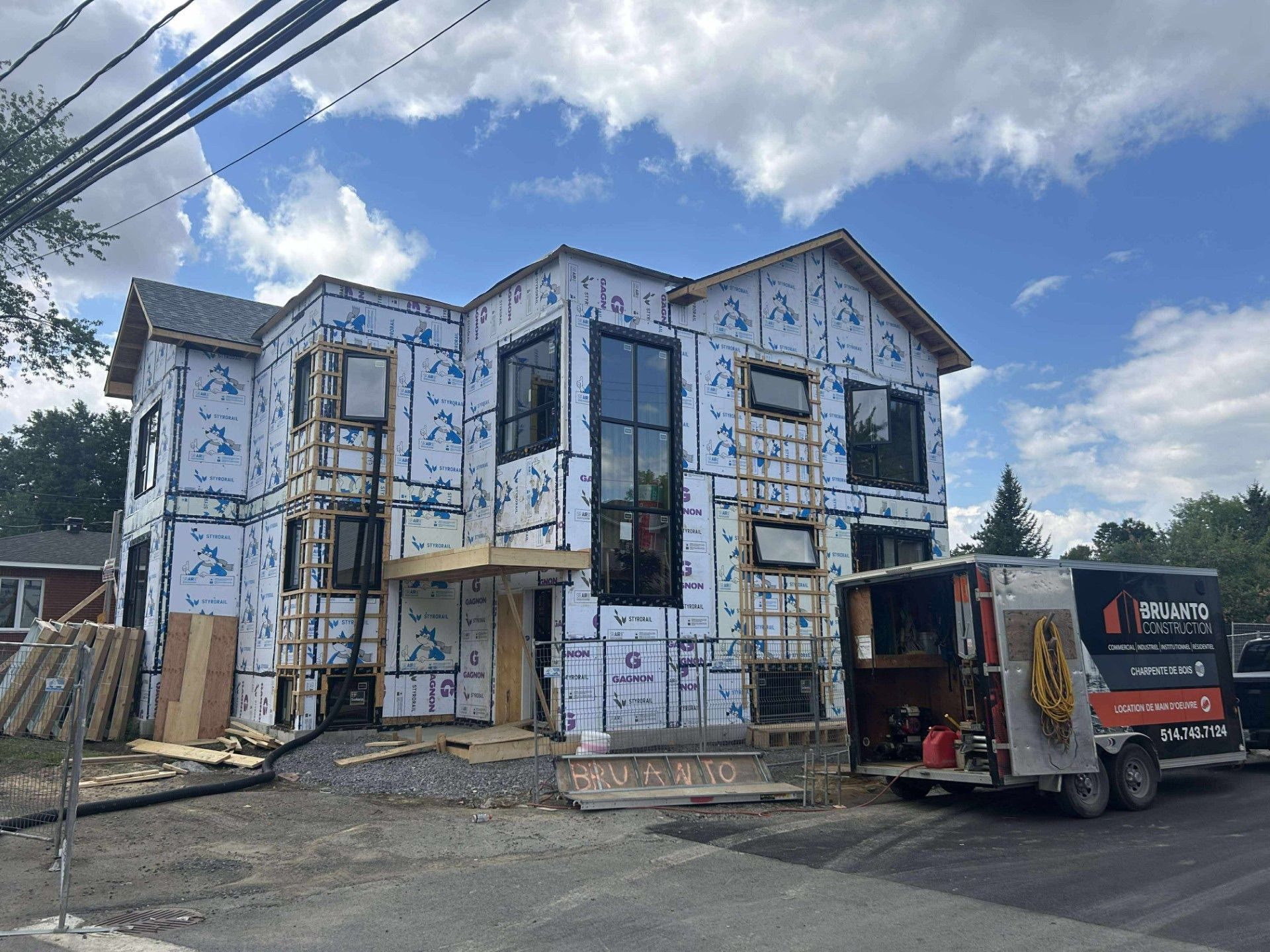Maison à deux étages en construction, avec un revêtement bleu, des fenêtres noires et une roulotte de chantier sur la droite.