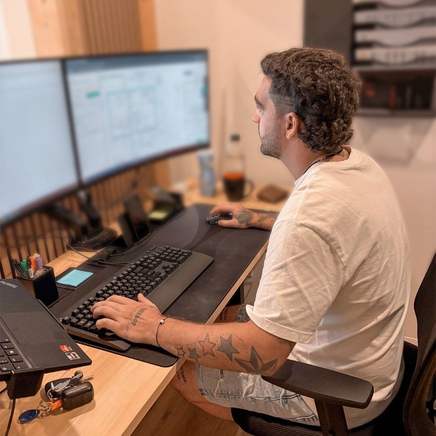 Un homme assis à un bureau avec deux écrans d'ordinateur, tapant sur un clavier, avec des tatouages sur le bras.