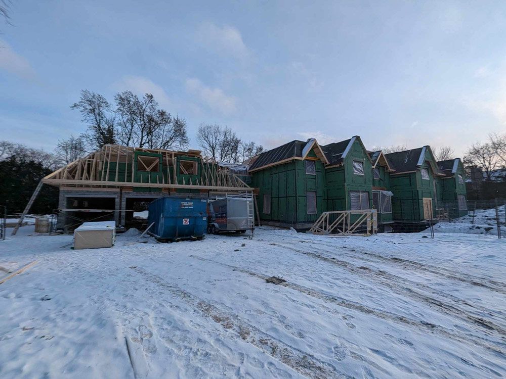 Chantier enneigé. Maisons et garage inachevés à ossature bois, benne à ordures bleue.