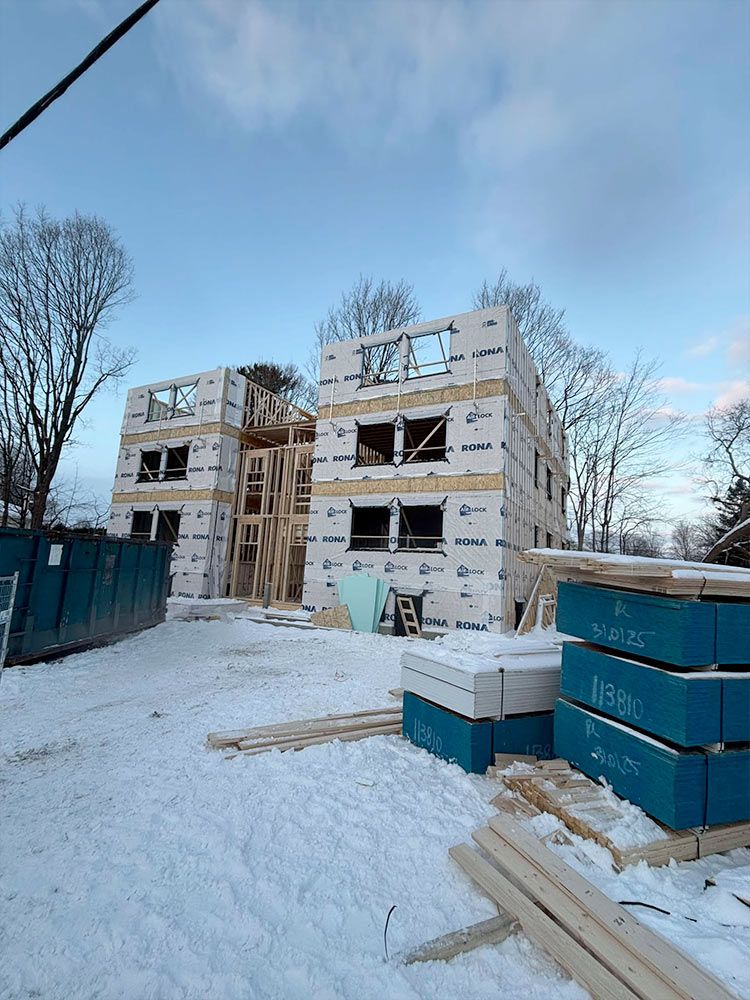 Bâtiment de deux étages en construction, recouvert de bâches blanches. Échafaudages en bois, bennes bleues et bois de construction sur un sol enneigé.