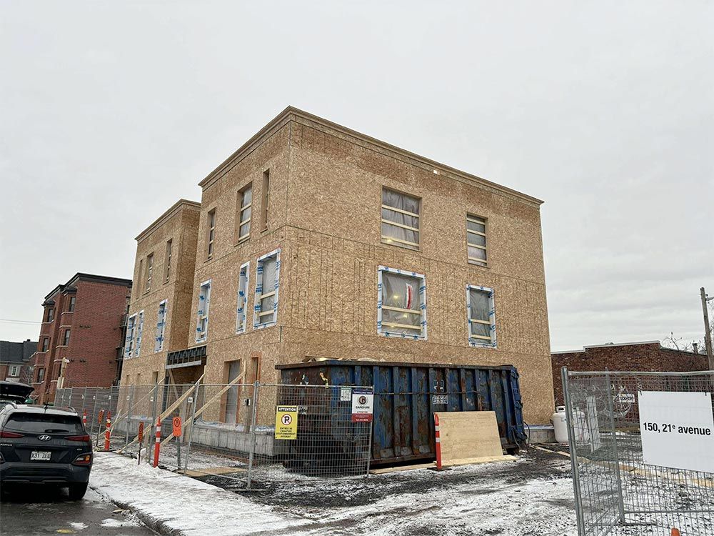 Bâtiment en construction avec ossature bois, fenêtres et benne à ordures, entouré d'une clôture de chantier.